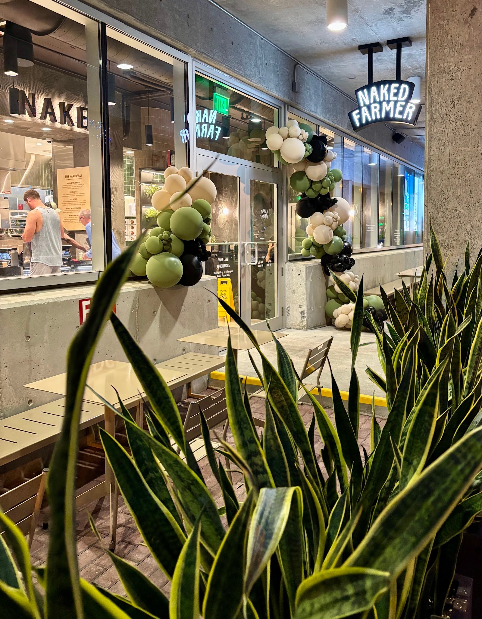 Storefront with a glowing Naked Farmer sign, glass windows, balloon garlands, outdoor tables, and tall plants in front.
