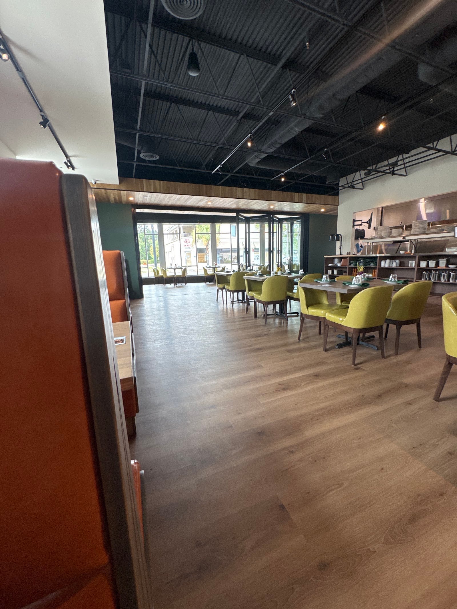 Empty restaurant dining room with green chairs, wooden tables, large front windows, and a dark exposed ceiling.